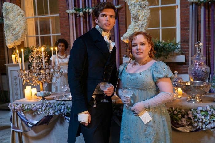 Luke Newton and Nicola Coughlan at an elegant event, in period attire, holding glasses. Newton wears a dark suit; Coughlan is in a blue gown. Candles and flowers adorn the background