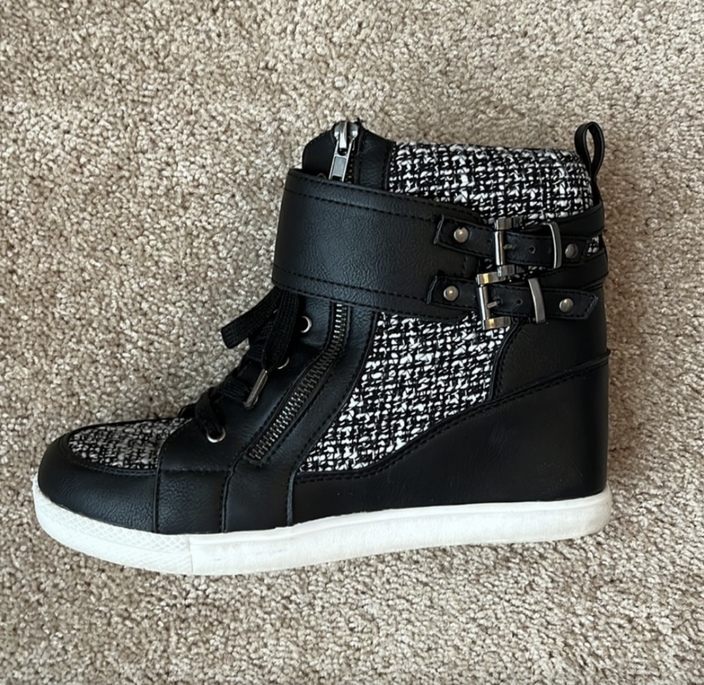 A stylish sneaker with a high-top design featuring black leather accents, textured fabric panels, double buckle straps, and a side zipper on a carpeted background