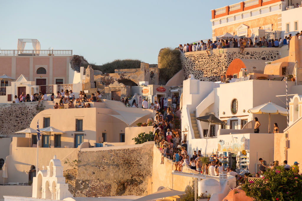 People gather at a crowded viewpoint in Santorini, Greece, atop whitewashed buildings and narrow streets, capturing the scenic sunset