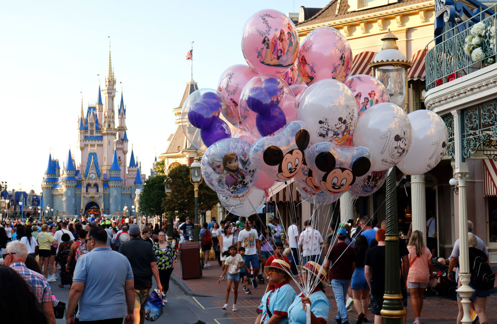 People walk down a busy street at a Disney theme park. A vendor holds colorful character balloons featuring Mickey Mouse and other Disney characters