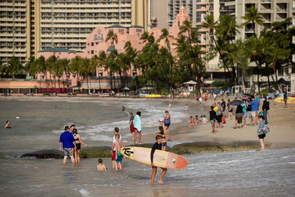 People enjoying a sunny day at a crowded beach with a surfer holding a surfboard and hotels visible in the background
