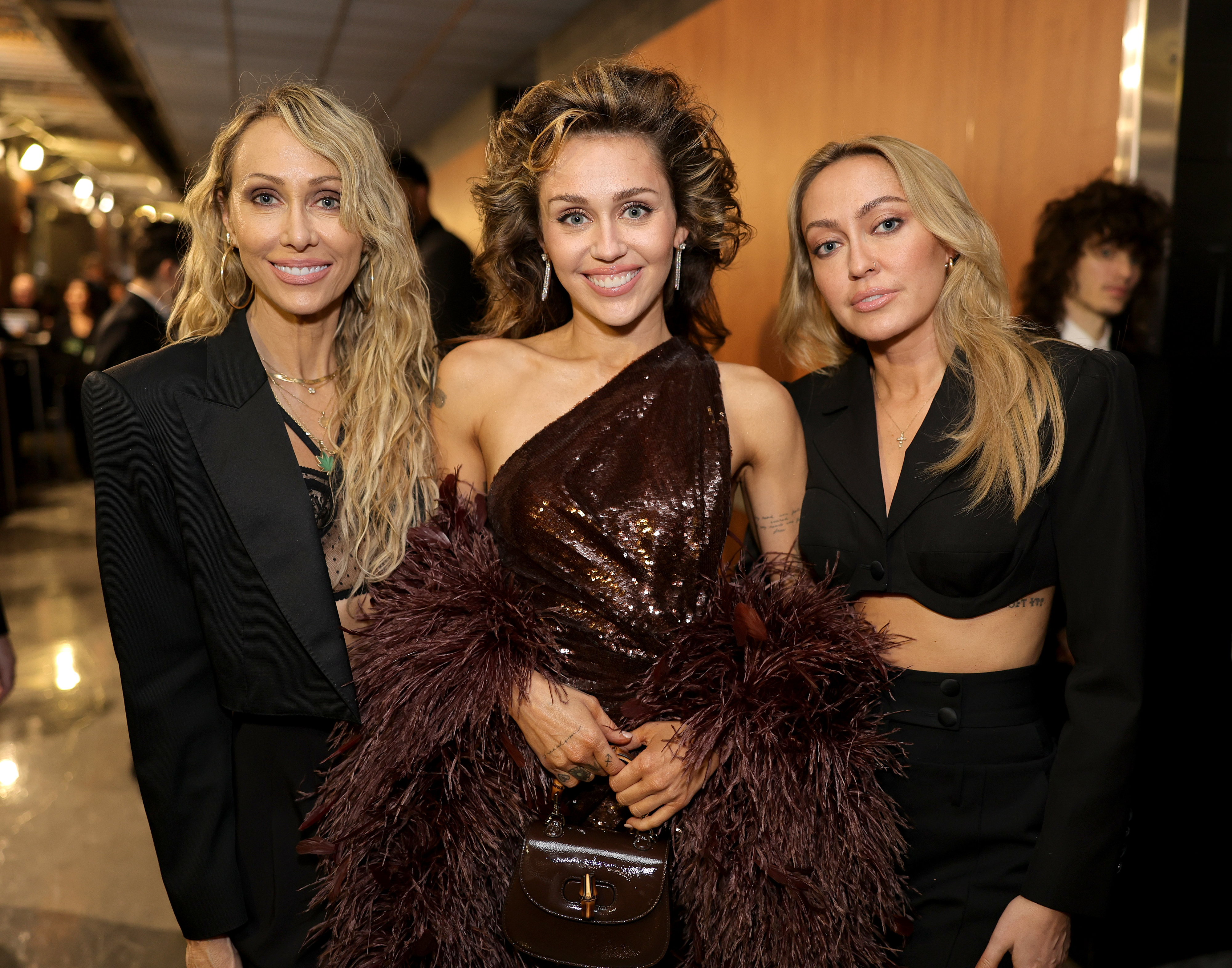 Tish Cyrus, Miley Cyrus, and Brandi Cyrus at a public event. Miley is wearing a one-shoulder sparkling dress with feather details, while Tish and Brandi are in black outfits