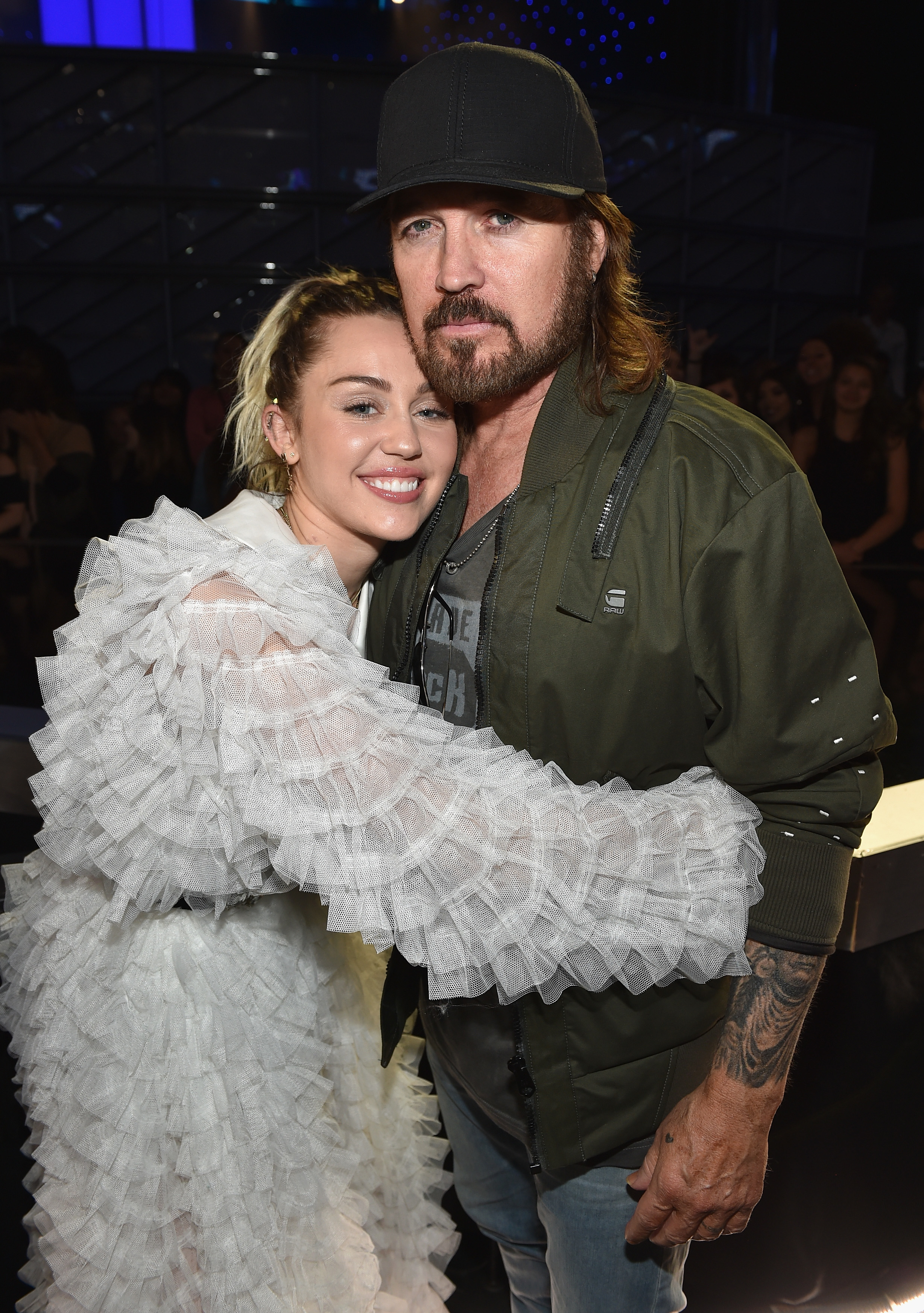 Miley Cyrus, in a ruffled outfit, embraces Billy Ray Cyrus, who wears a cap and jacket, at a celebrity event