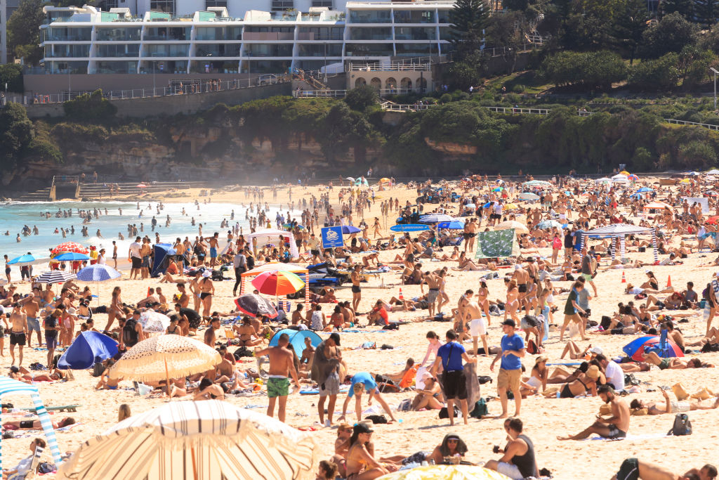 A crowded beach scene with many people sunbathing, swimming, and enjoying the day. In the background, modern buildings are visible