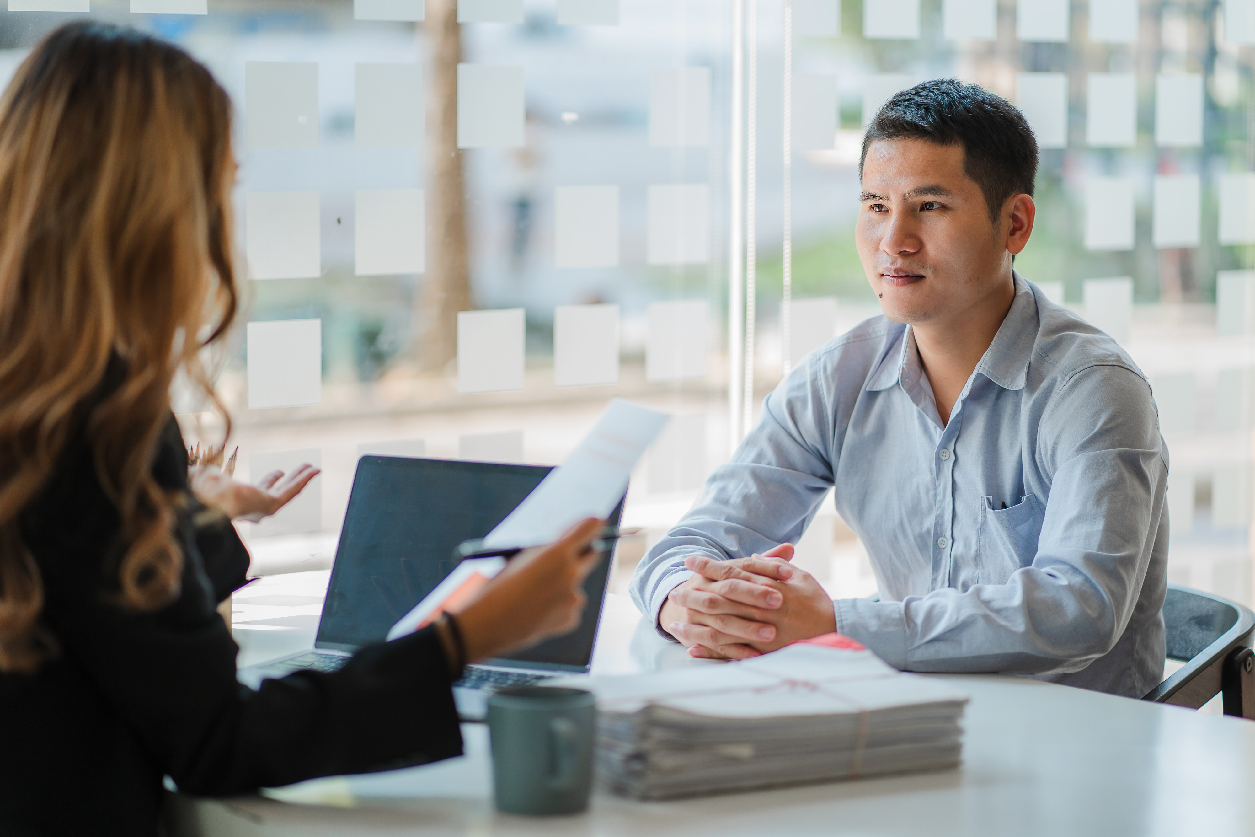 A woman interviews a man at a desk with documents and a laptop in a professional setting