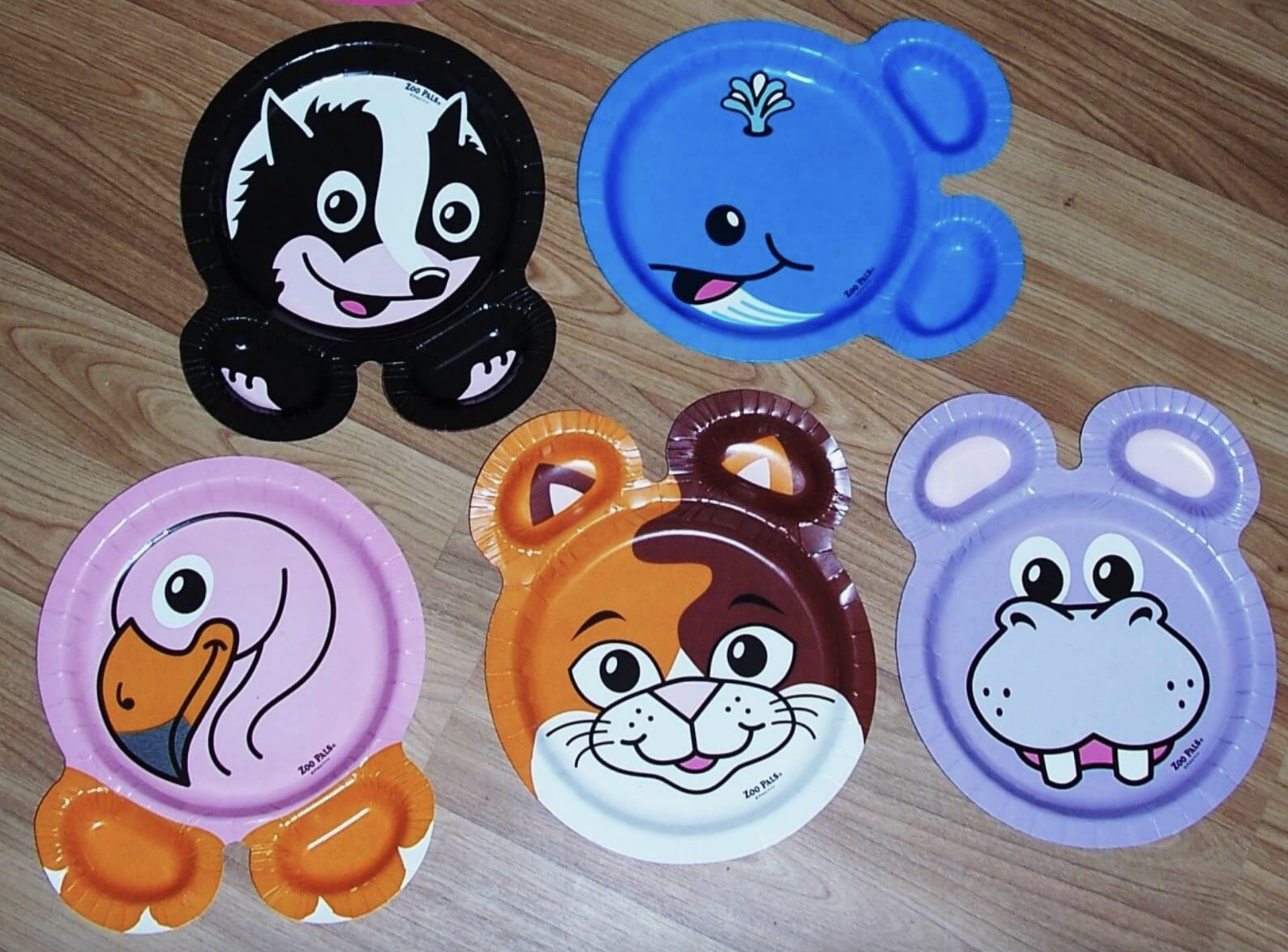 Five cartoon animal-themed Zoo Pals paper plates on a wooden surface; the plates feature a raccoon, whale, flamingo, cat, and hippo