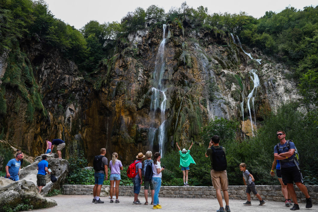 A group of people stands at the base of a rocky waterfall, some taking photos and others climbing nearby rocks. An adult lifts a child beside the waterfall