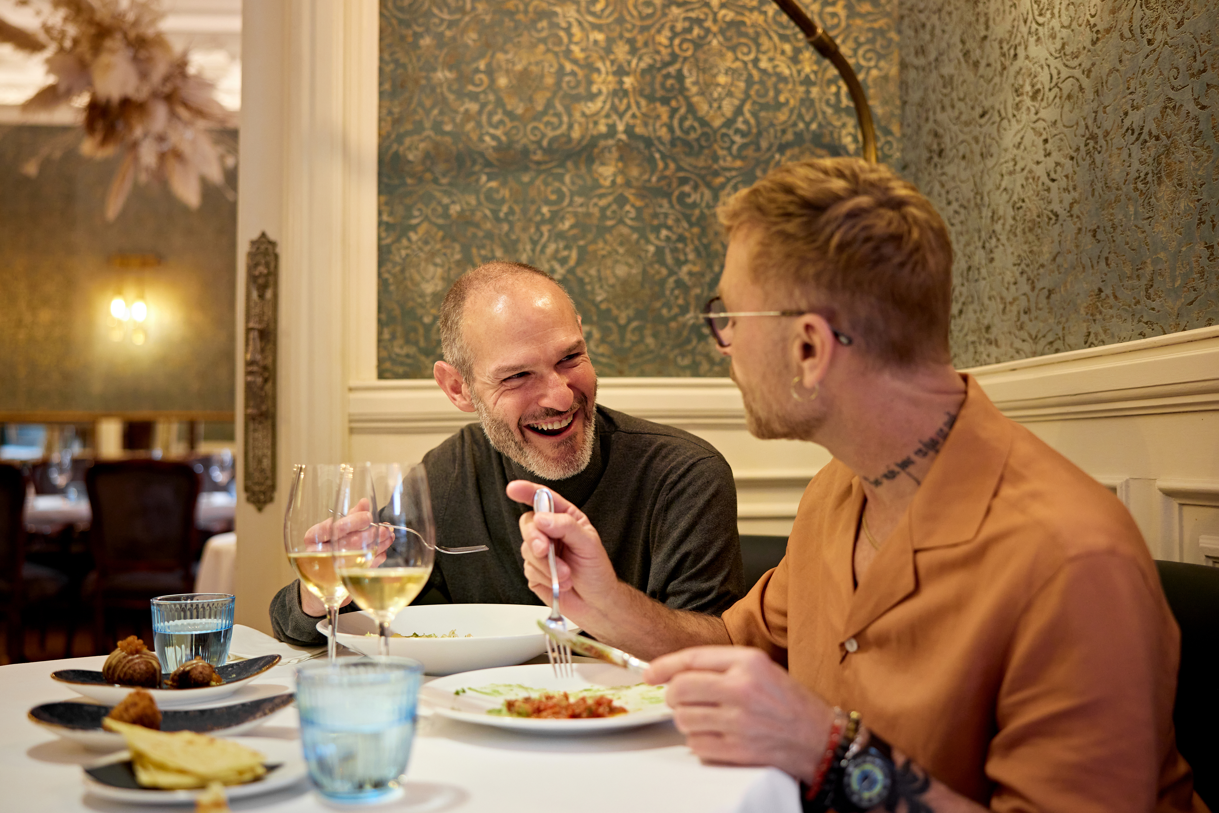 Two men having a lively conversation while dining at an elegant restaurant. One man wears a gray sweater, and the other wears a brown shirt with rolled-up sleeves