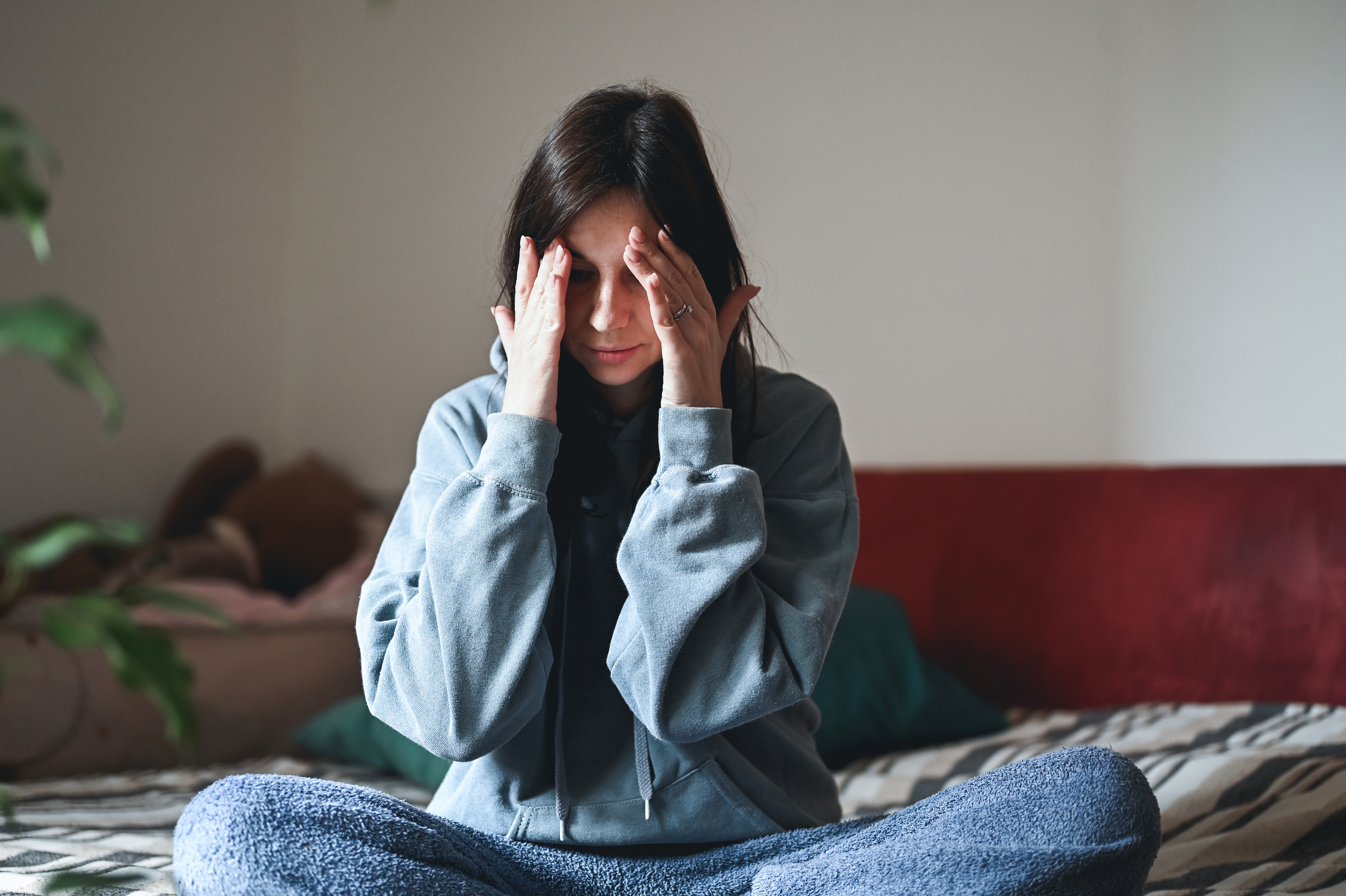 Woman wearing a sweatshirt and loose pants, sitting cross-legged on a bed, with hands on her head, looking stressed or in deep thought