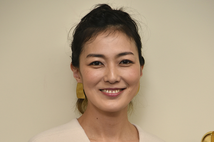女性がベージュのシンプルなドレスを着て微笑んでいる。背景はクリーム色の壁。