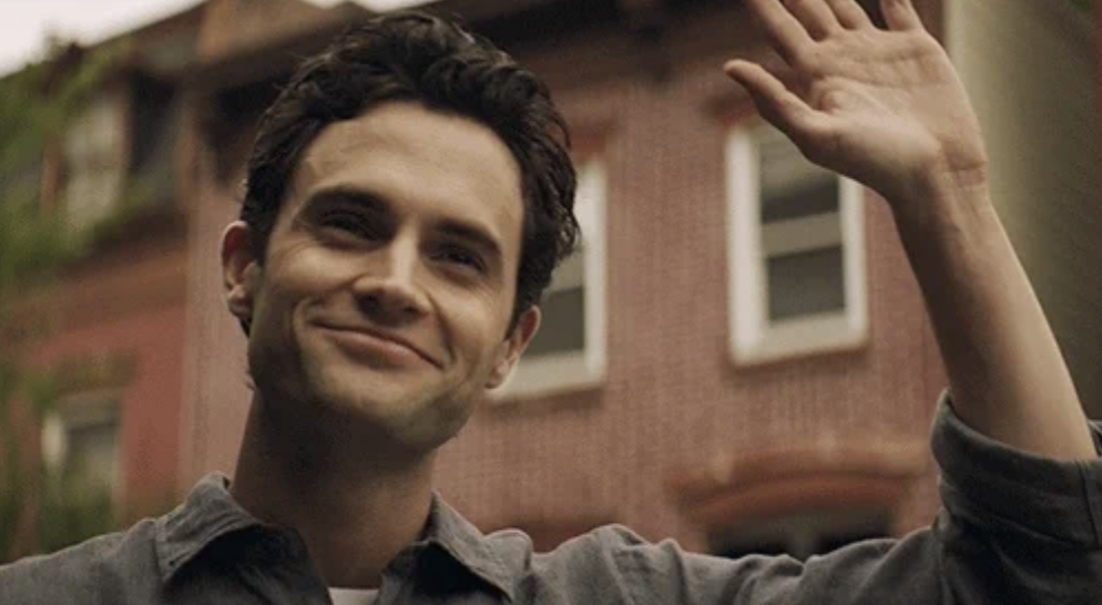 Penn Badgley, smiling and waving, stands in front of a brick building
