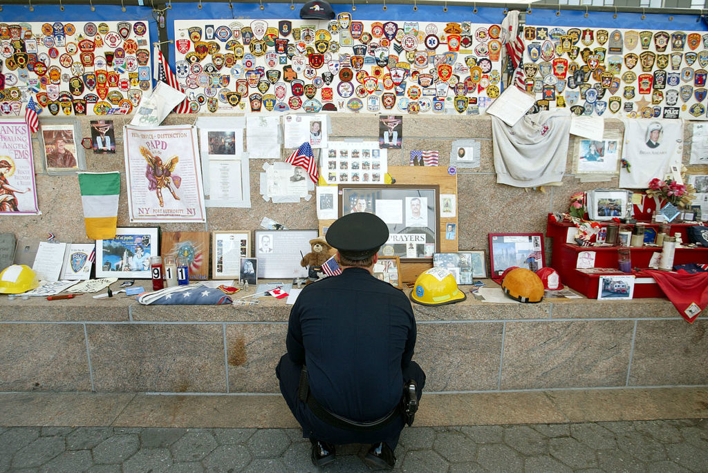 A police officer kneels in front of a 9/11 memorial wall covered with photos, badges, letters, and various memorabilia, honoring the victims of the terrorist attacks