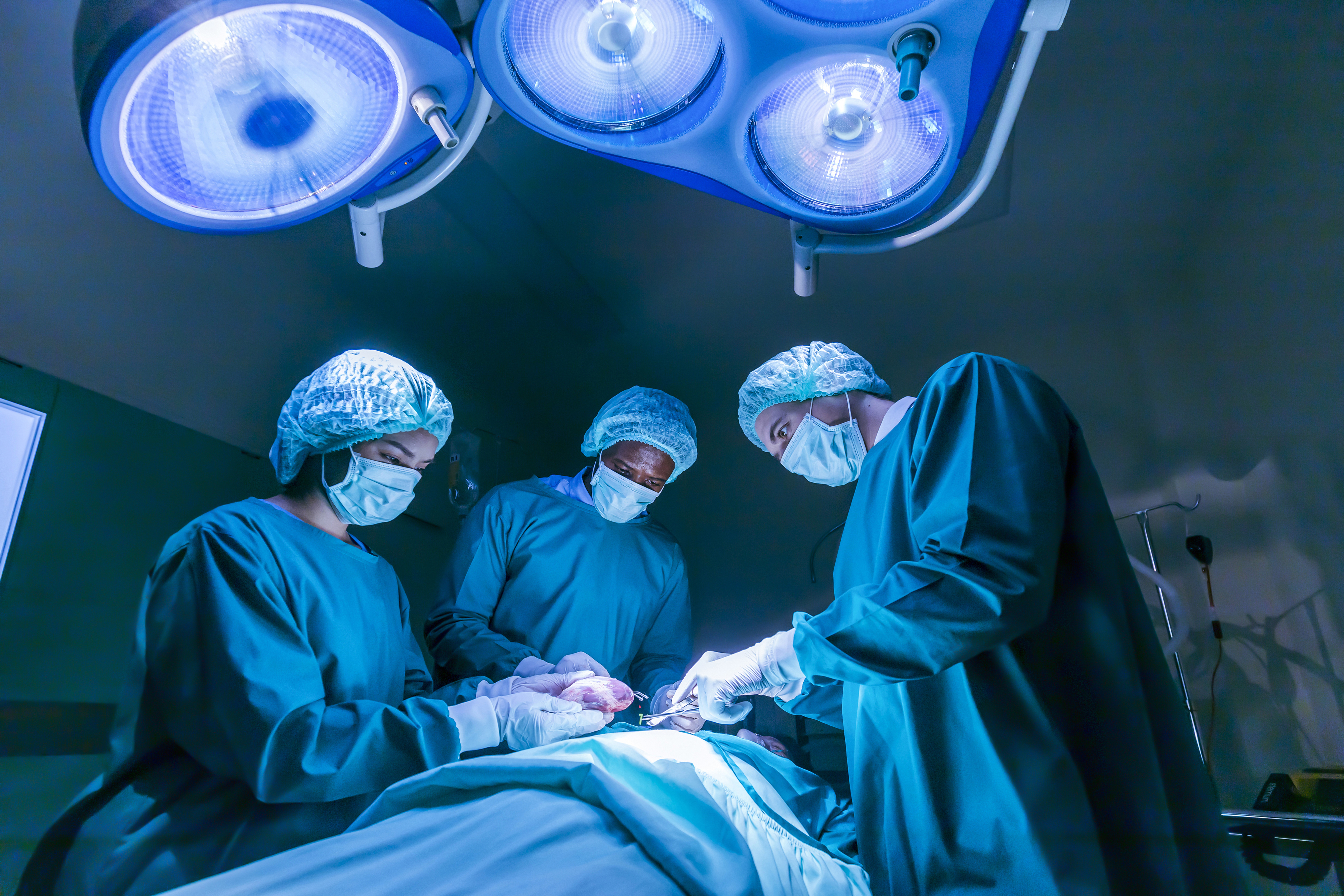 Surgeons in an operating room performing surgery under bright surgical lights, wearing scrubs, caps, and masks