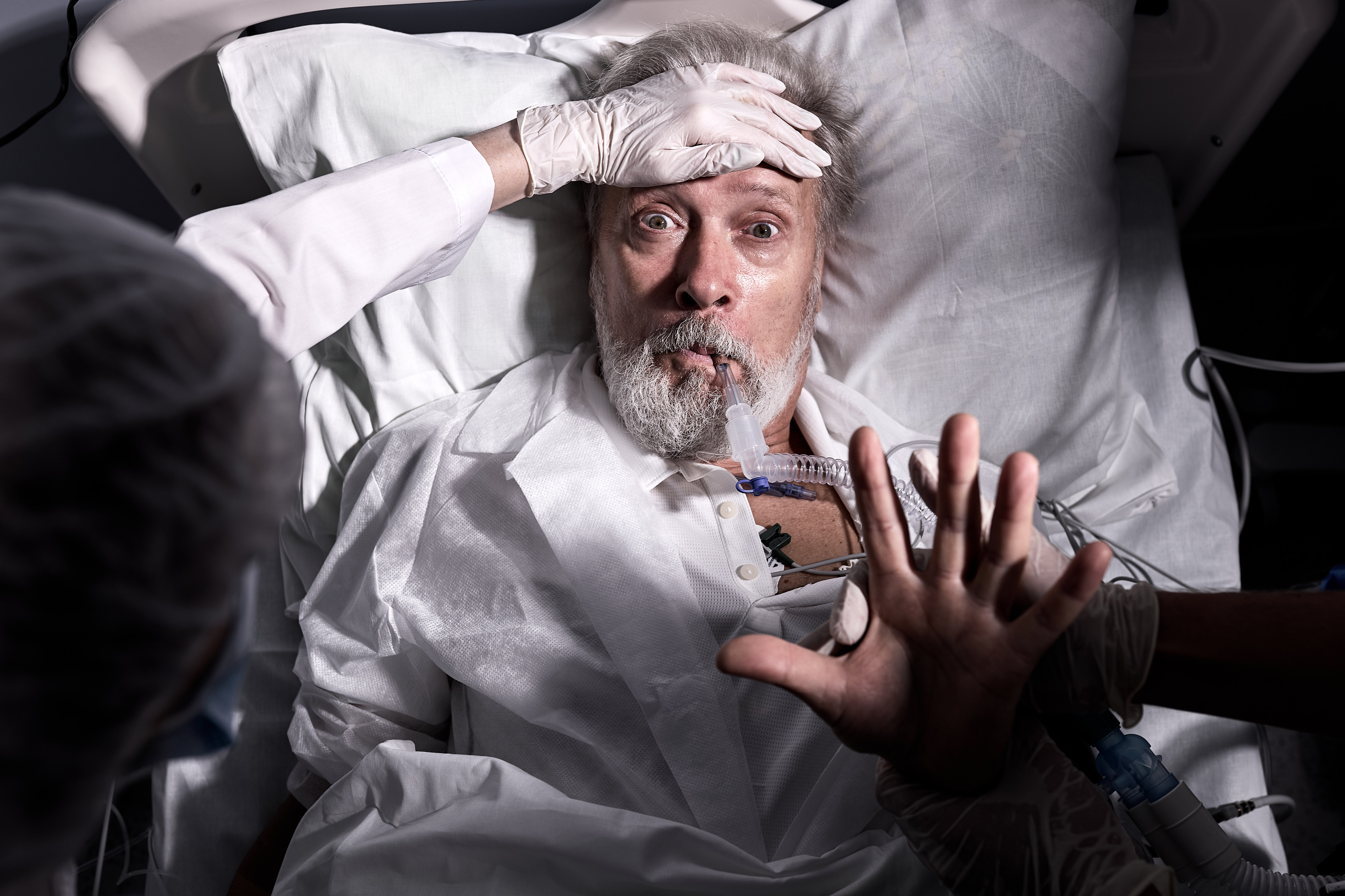 An older man with a surprised expression lies in a hospital bed, wearing a medical gown and connected to a ventilator. A medical professional&#x27;s gloved hands are on his forehead and near his arm