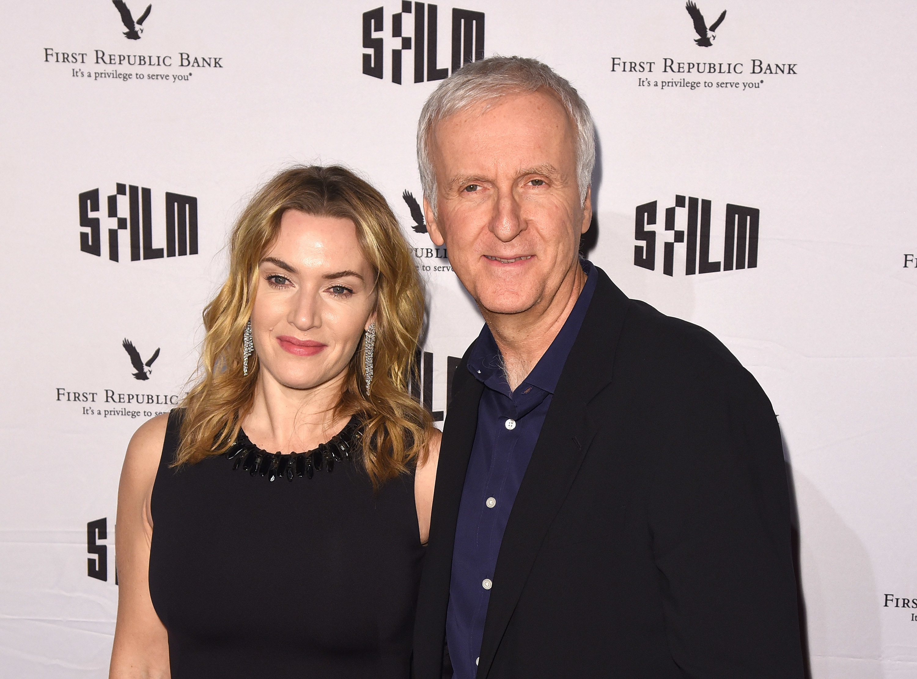 Kate Winslet and James Cameron pose together at a SFILM event backdrop. Kate wears a sleeveless dress with a beaded neckline, and James is in a dark blazer