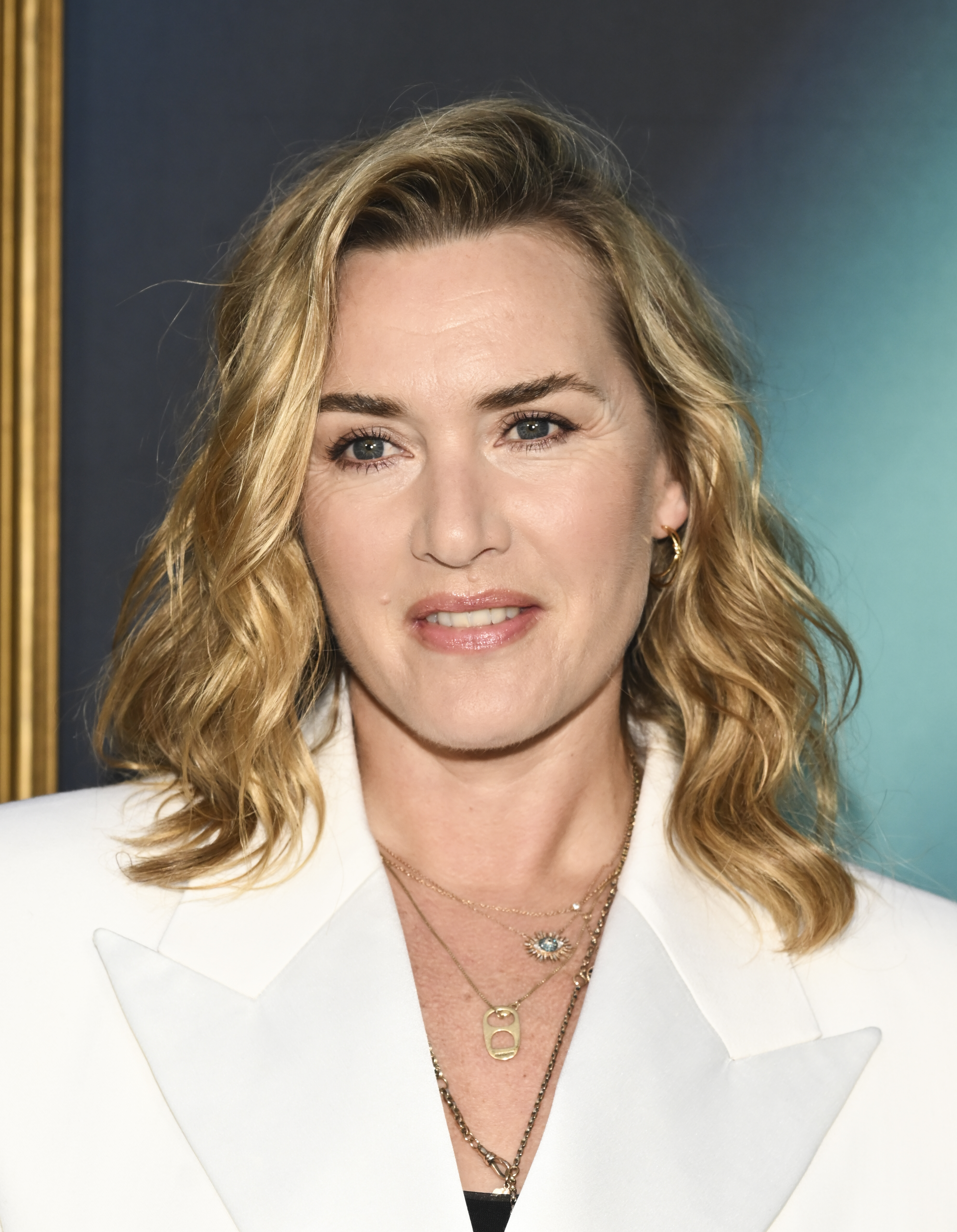 Kate Winslet in a blazer, layered necklaces, and wavy hair at a formal event. She is smiling softly and facing the camera