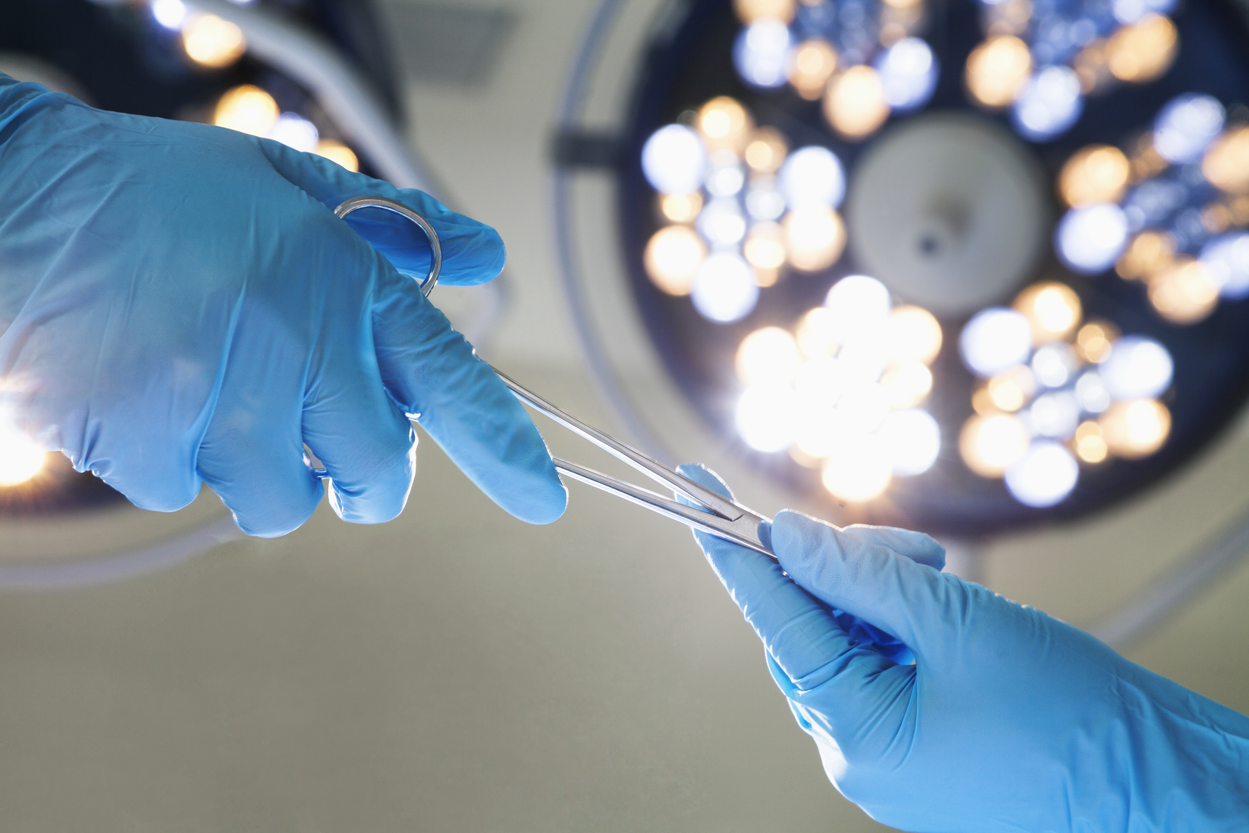 A surgeon in blue gloves hands a medical instrument to another surgeon under bright operating room lights