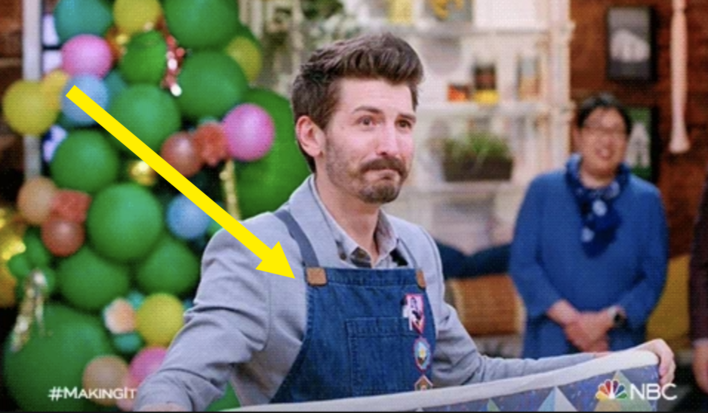 Nick Offerman on NBC's &quot;Making It,&quot; wearing a light gray shirt and overalls, smiling. In the background, a person in blue stands near colorful decorations