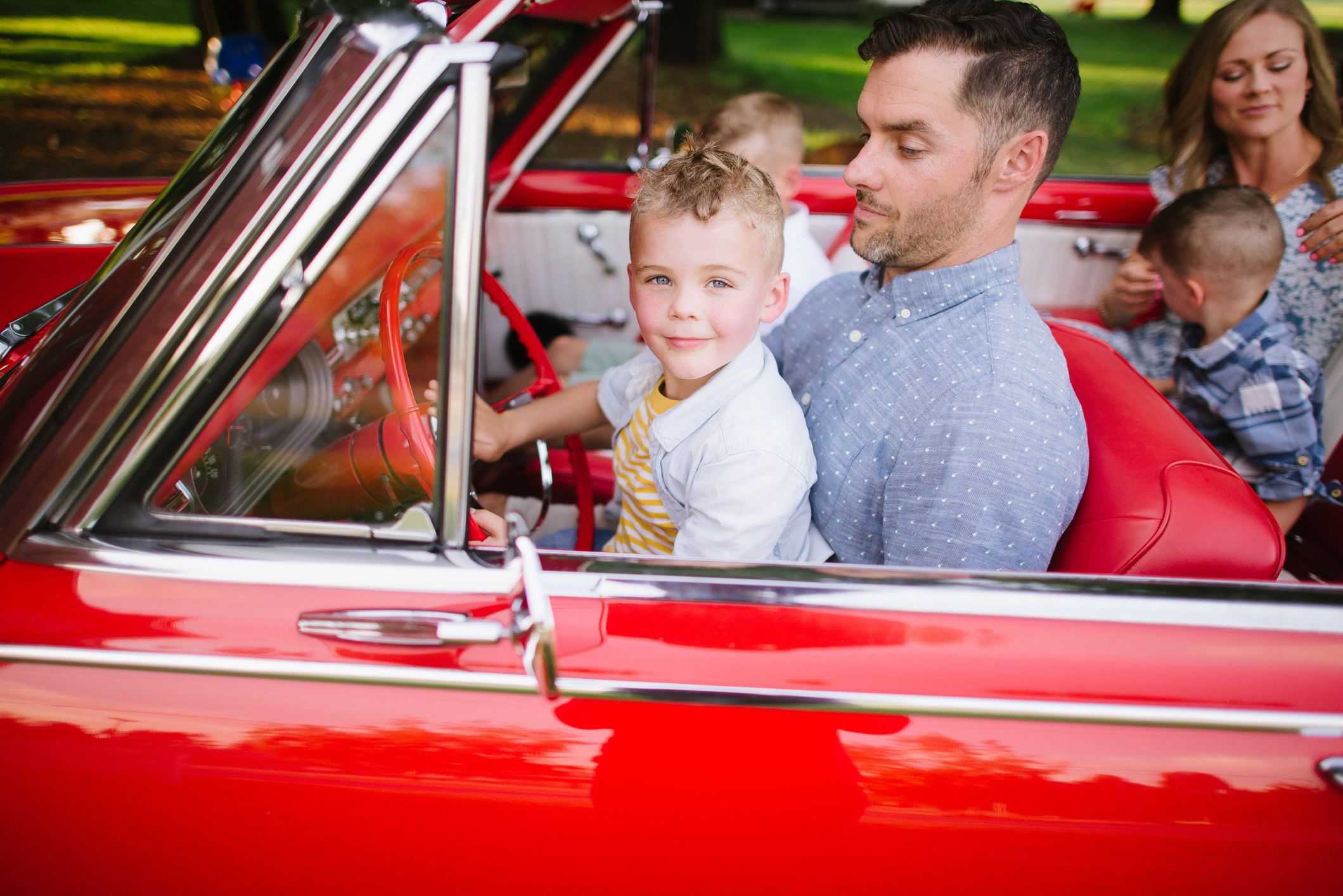 A man drives a vintage car with a young boy in his lap. Another woman and two children are in the backseats