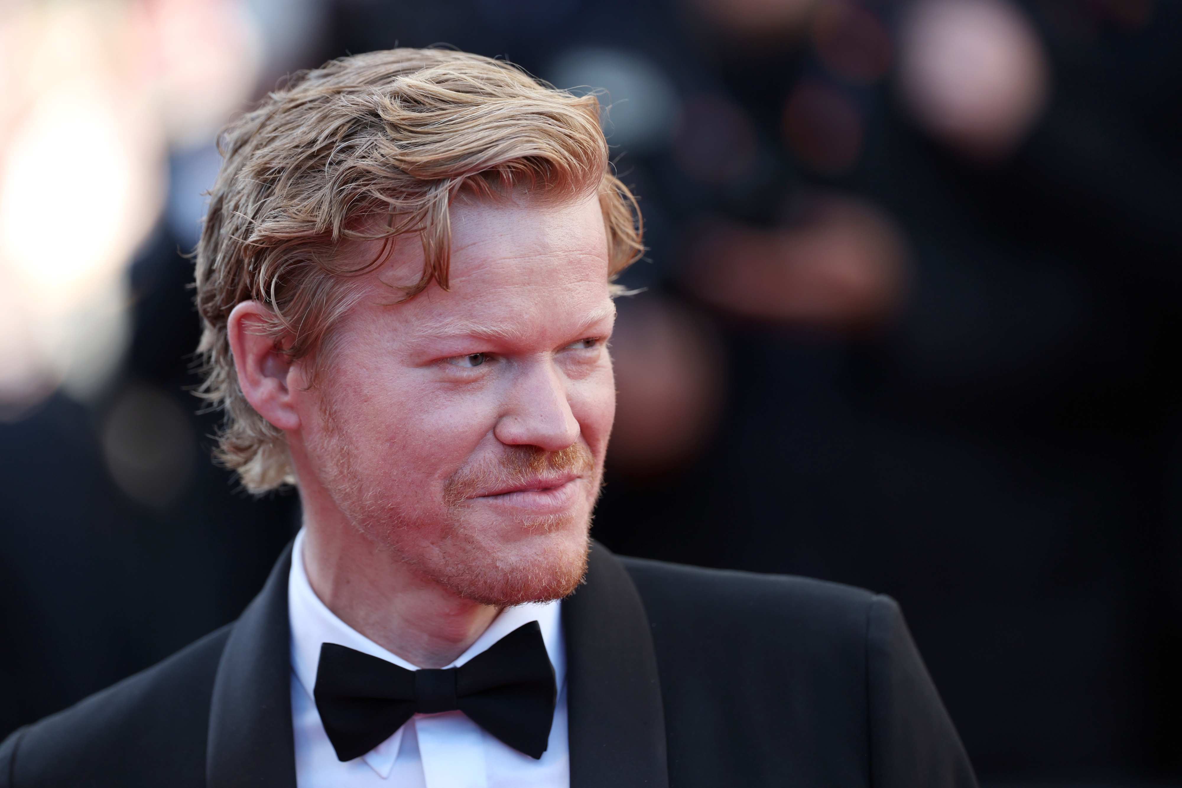 Jesse Plemons wearing a black tuxedo with a bow tie, smiling on a red carpet