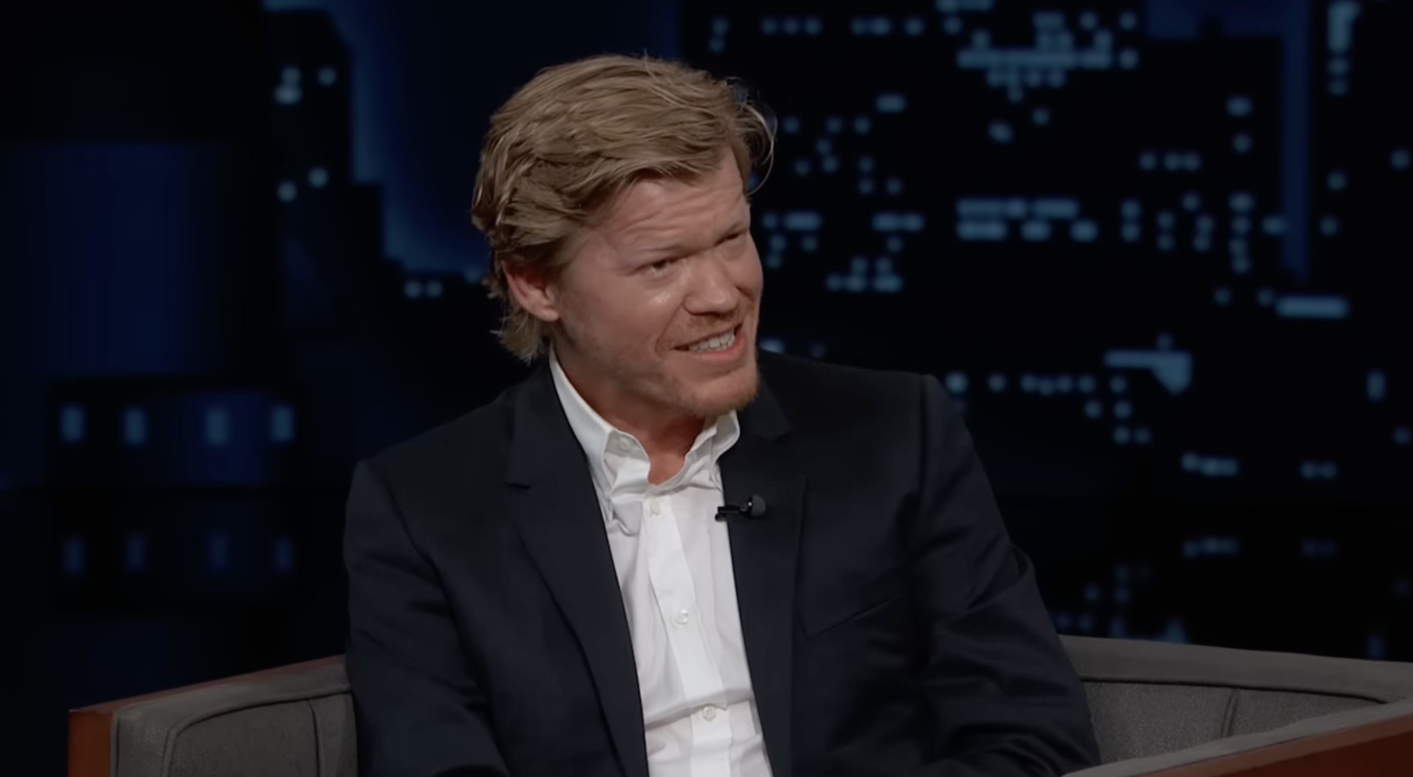 Jesse Plemons on a talk show set, seated and smiling during an interview, wearing a dark suit with an open-collared shirt