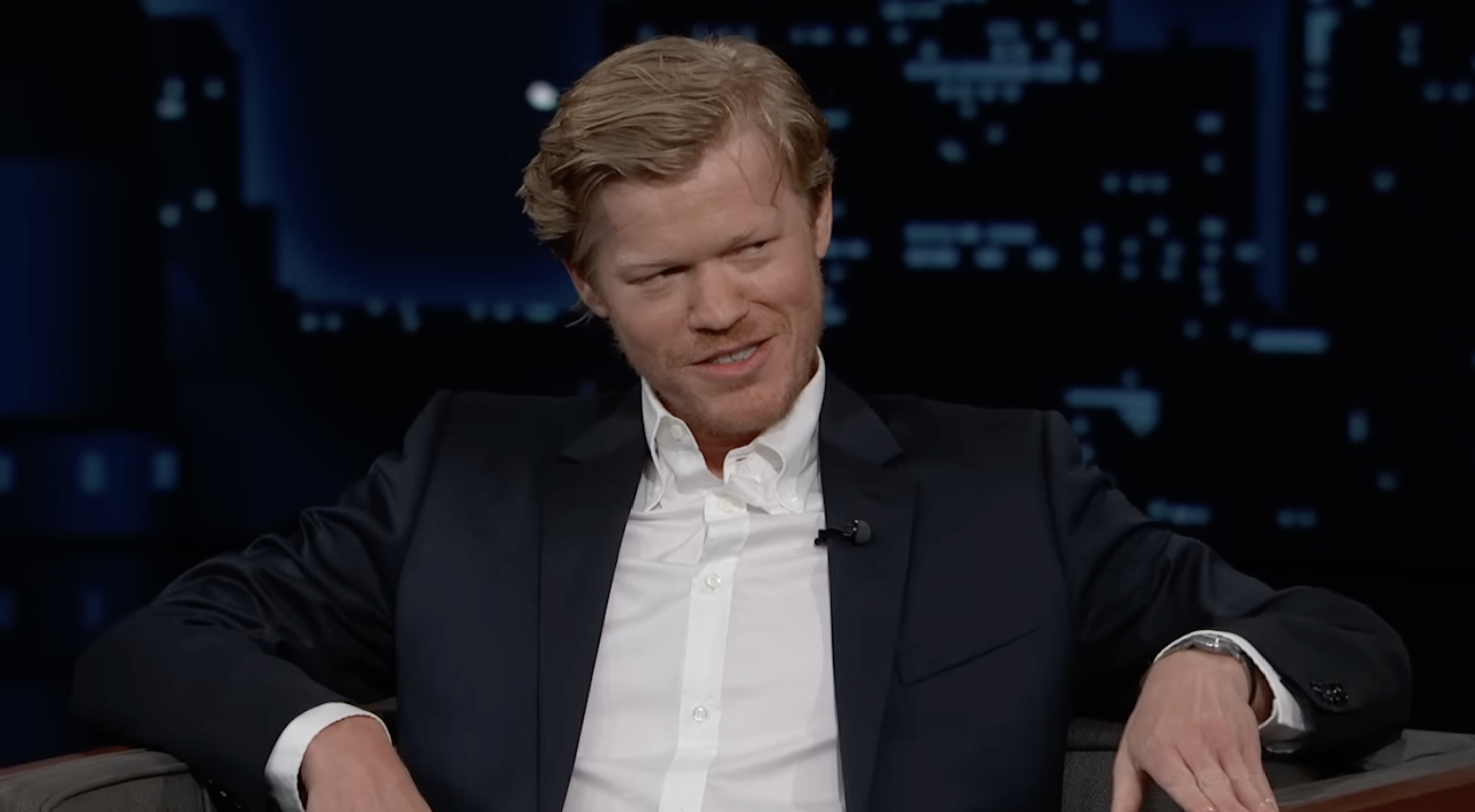 Jesse Plemons wearing a suit and white shirt, sitting and speaking on a late-night talk show set