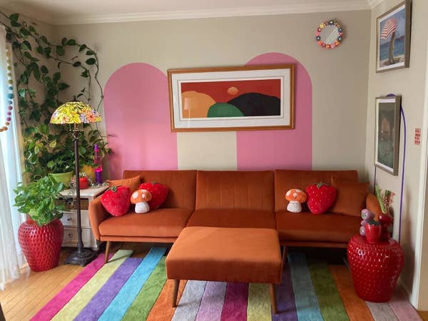 A reviewer&#x27;s living room scene with a modern, velvet sofa, vibrant mushroom and strawberry pillows, a colorful rainbow rug, plants, and a framed abstract painting