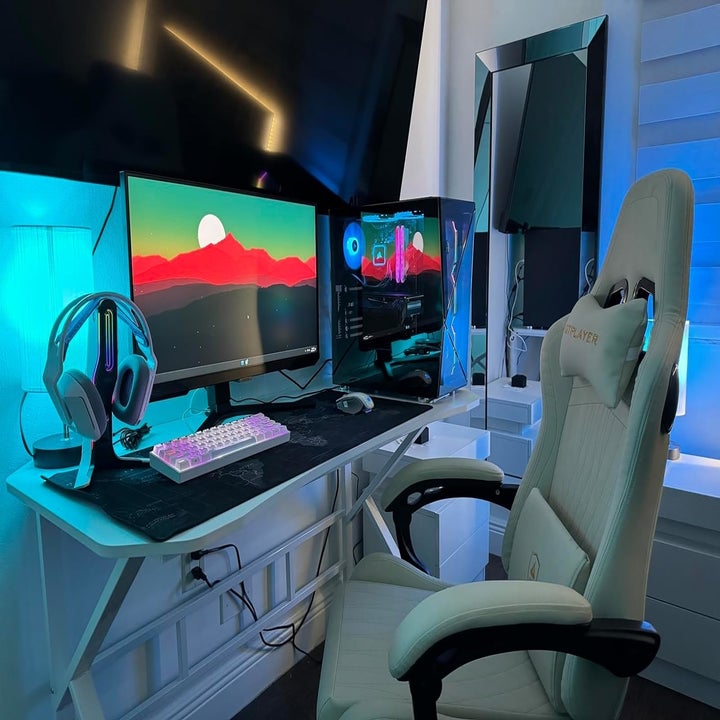 Gaming setup featuring a large monitor displaying a mountain landscape, gaming PC with LED lights, white gaming chair, headset, and multicolored keyboard