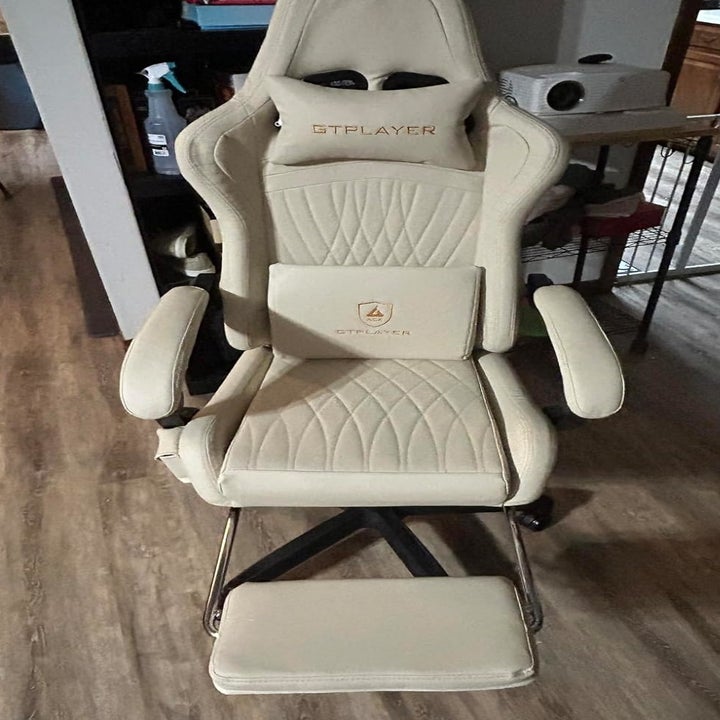 GTPLAYER beige office/gaming chair with armrests, headrest, and lumbar support cushion, positioned in a home setting with wooden floor and kitchen in the background