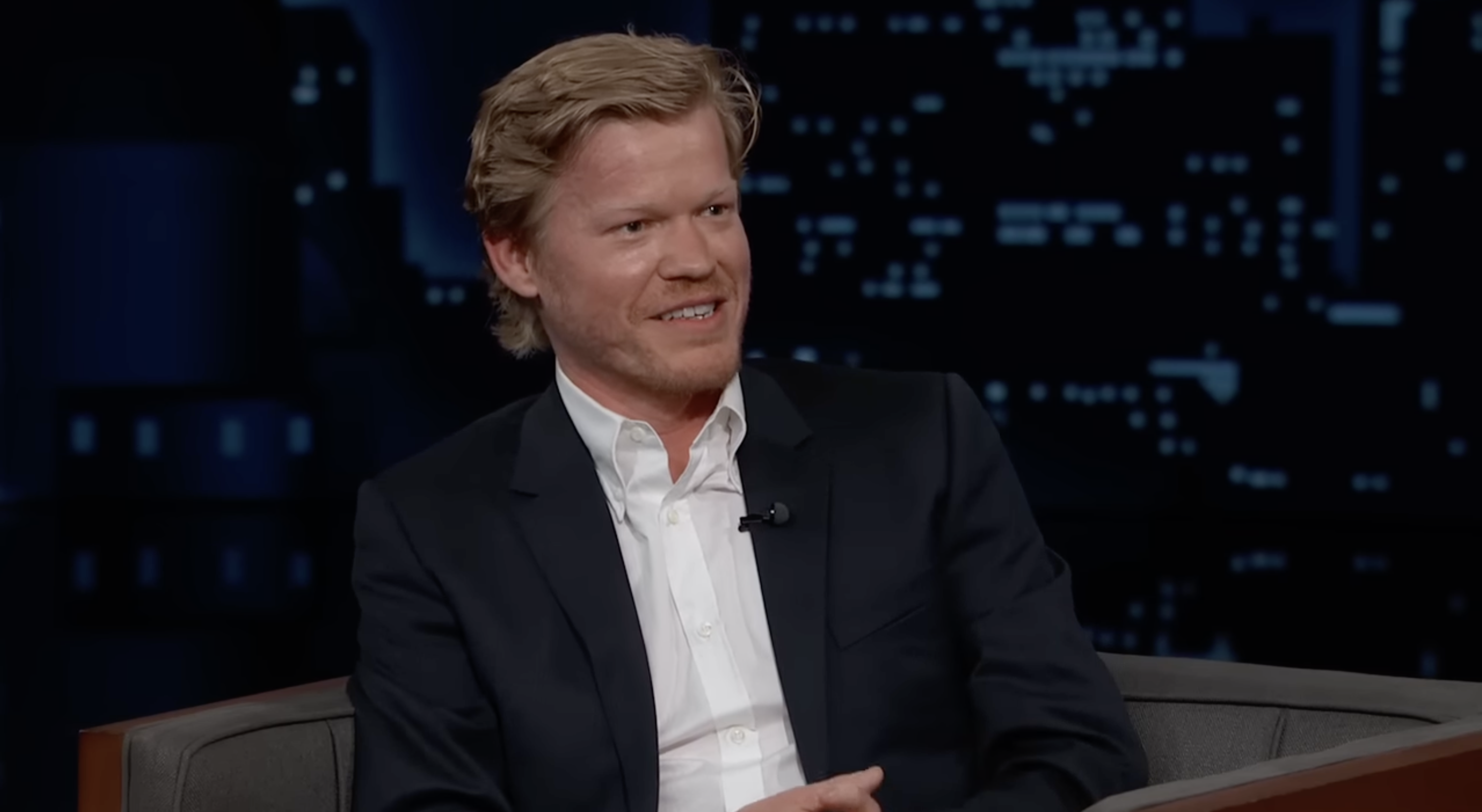 Jesse Plemons sits on a talk show set, wearing a white shirt and dark blazer