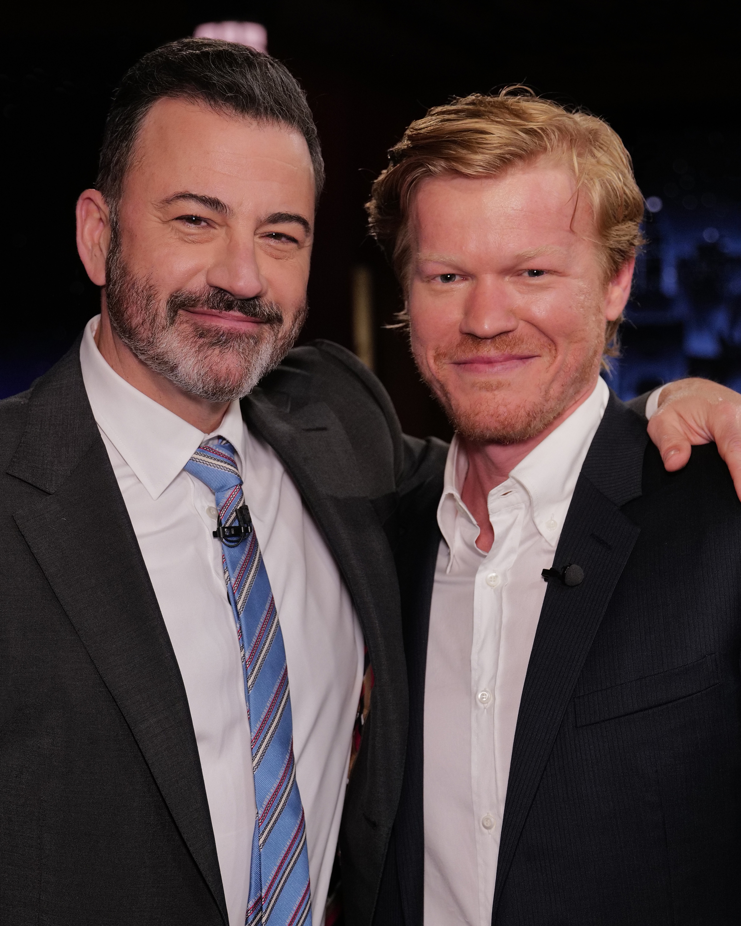 Jimmy Kimmel and Jesse Plemons, both in suits, pose together on a set. Kimmel has his arm around Plemons