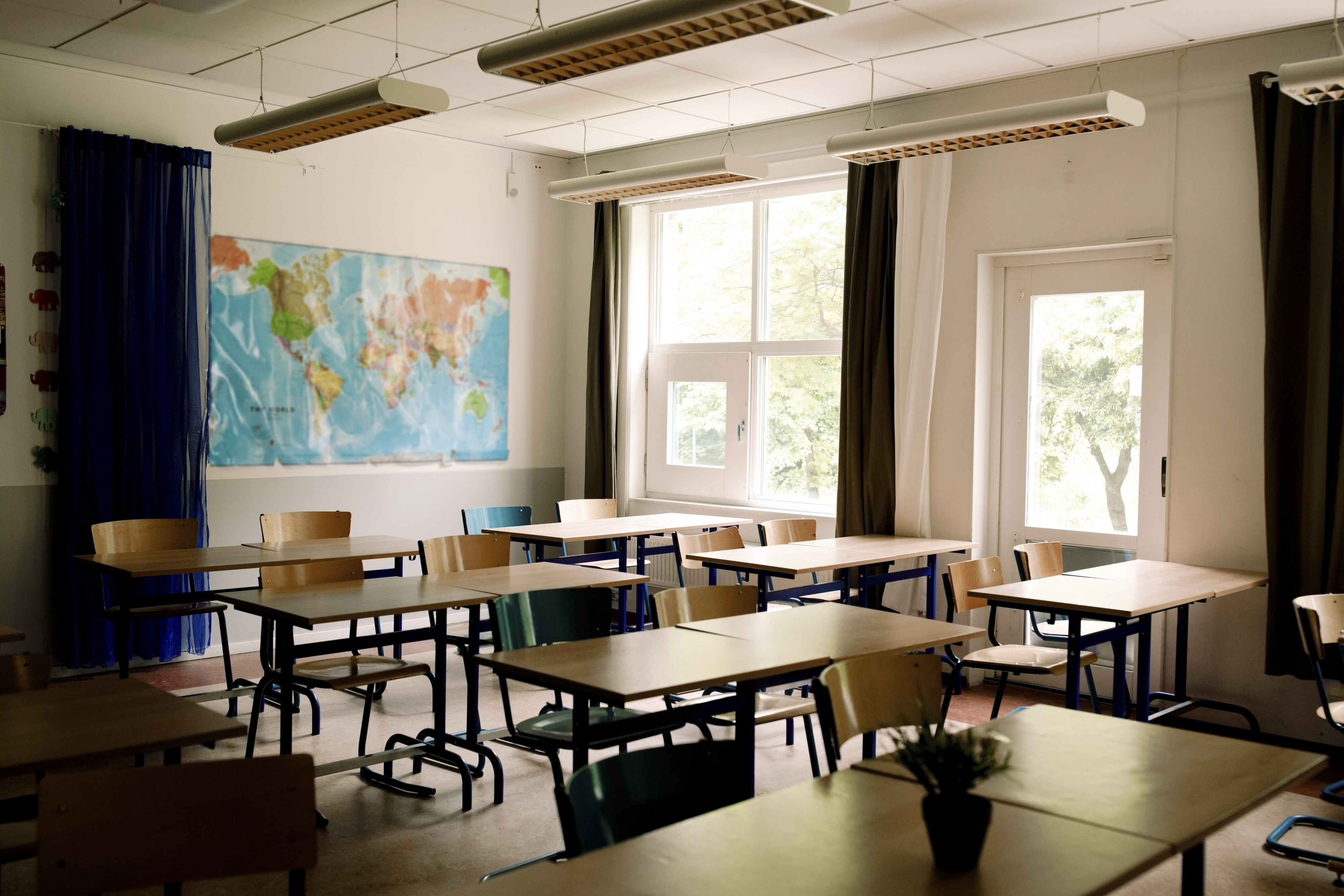 A classroom with multiple rows of desks and chairs, a world map on the wall, and large windows allowing natural light in