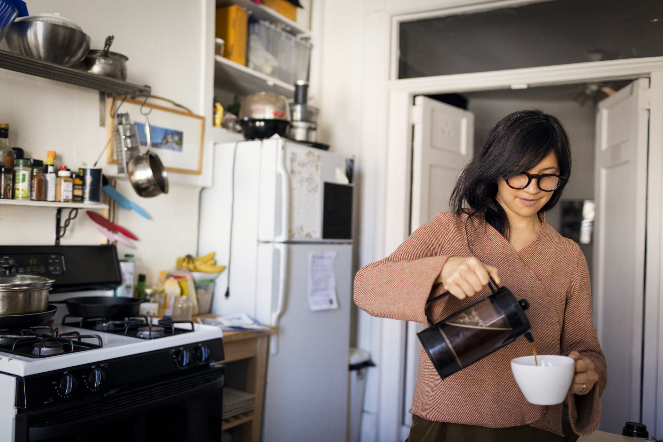 A person with glasses pours coffee into a mug in a cozy kitchen with various utensils and ingredients on shelves and counters