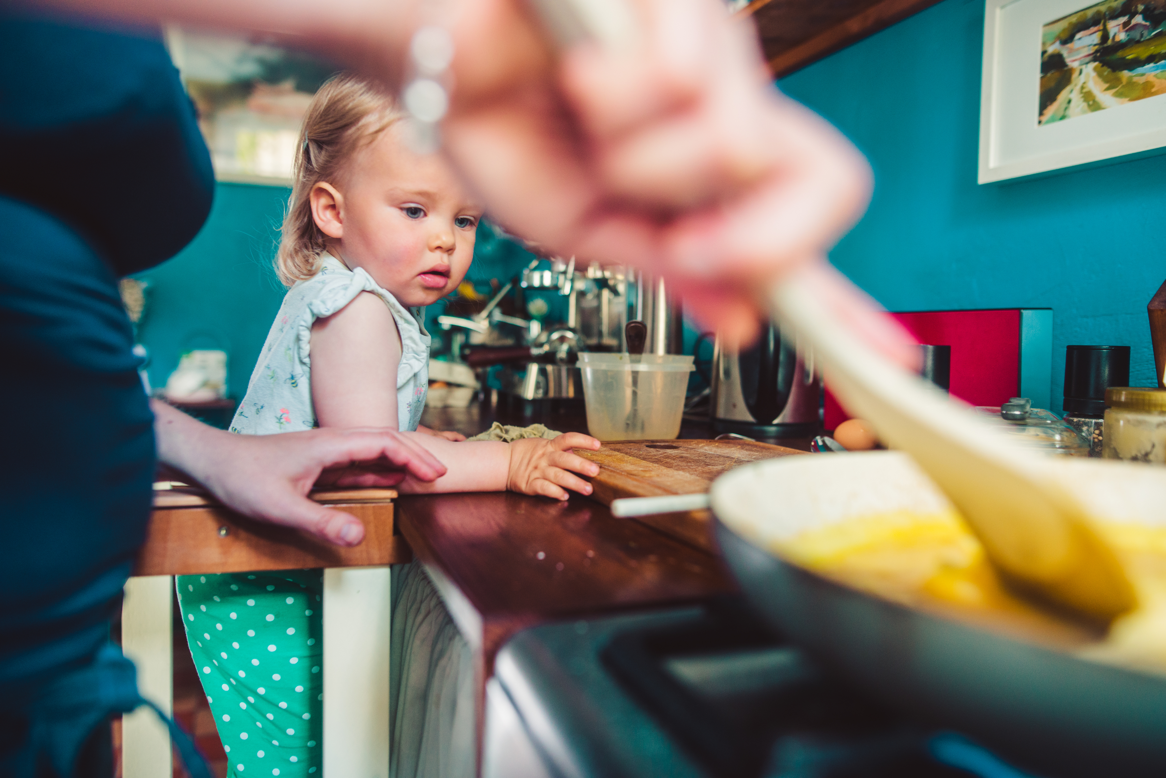 A toddler watches a woman stirring food in a frying pan in the kitchen. The toddler is wearing a light top and green pants with white dots