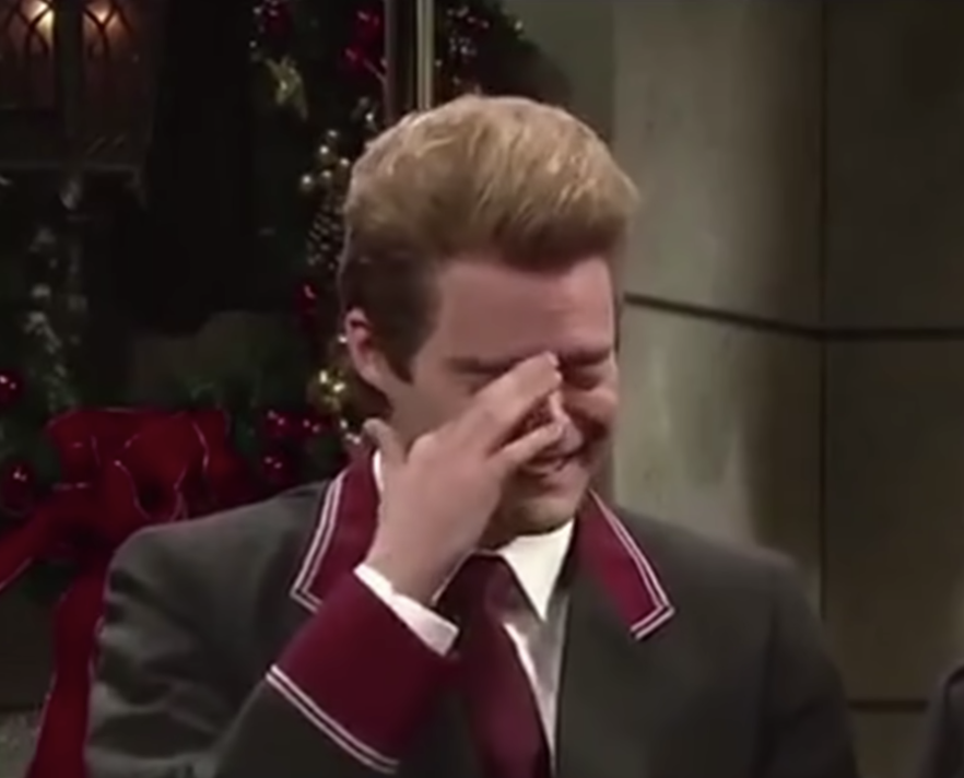 A man in a suit with ornamental trim laughs, covering part of his face with his hand, in what appears to be a festive, holiday-themed setting