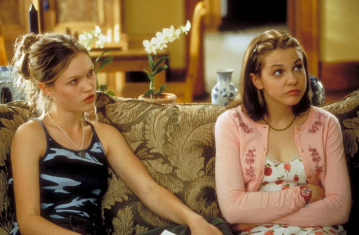 Julia Stiles and Larisa Oleynik sit on a couch, both wearing casual outfits, in a scene from "10 Things I Hate About You"
