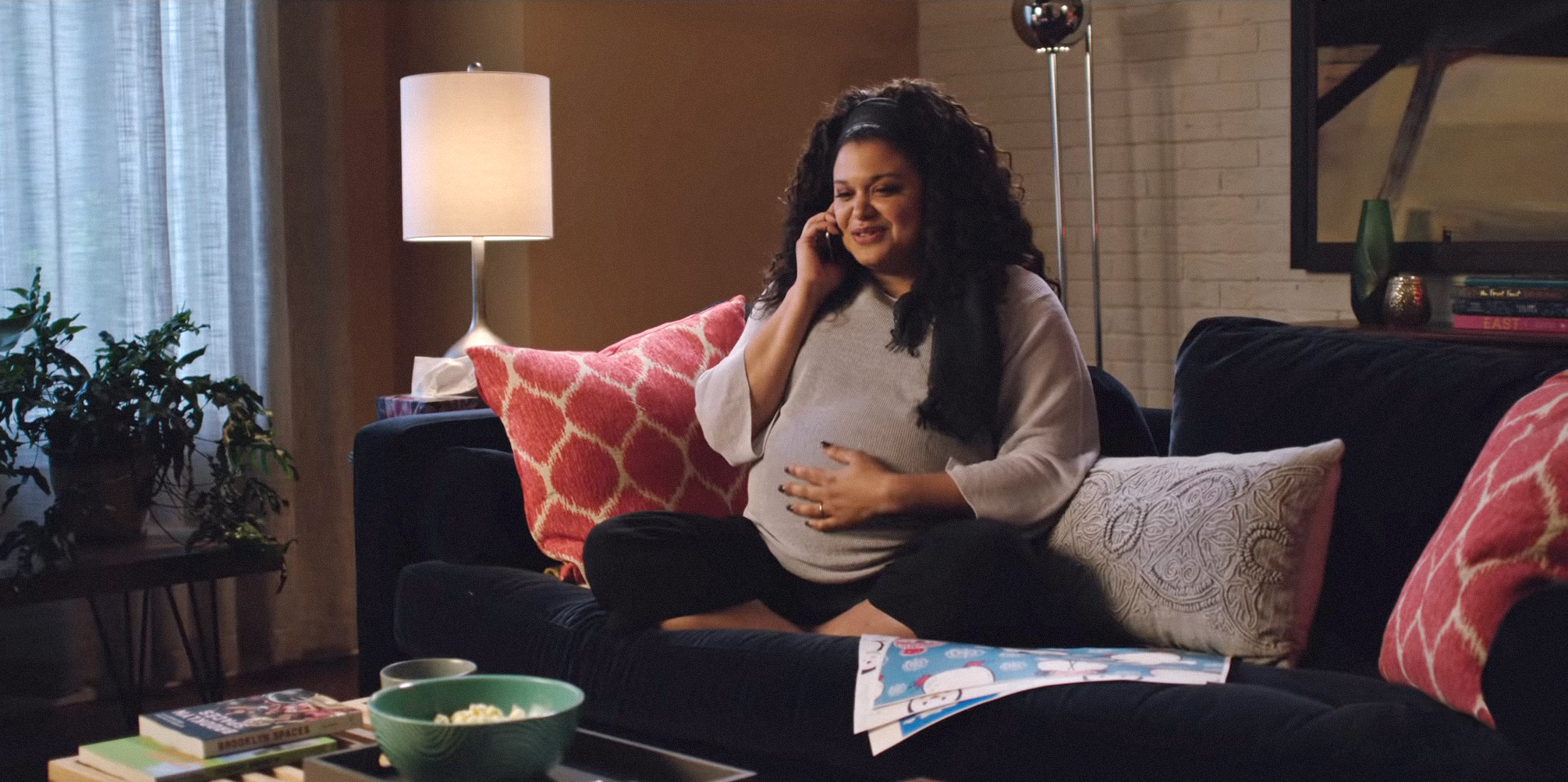 Michelle Buteau sitting on the couch, rubbing her pregnant belly while she talks on the phone