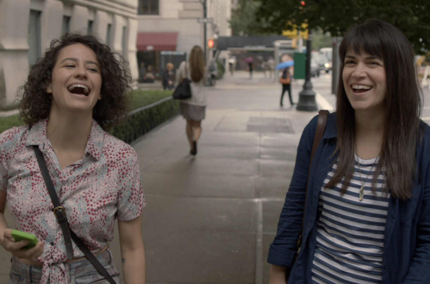 Ilana Glazer and Abbi Jacobson are walking down a city street, laughing joyfully. Ilana wears a patterned shirt, and Abbi wears a striped shirt and a jacket