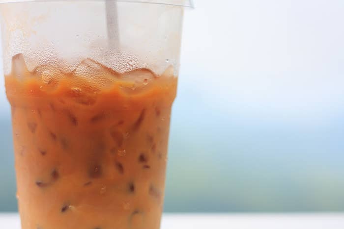 A close-up of a plastic cup filled with iced tea, including ice cubes and a plastic straw. The background is blurred