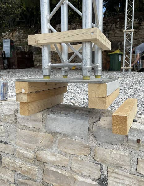 A makeshift stand is created using wooden blocks and a metal sheet to support a metal frame structure on a stone wall. No persons are present in the image