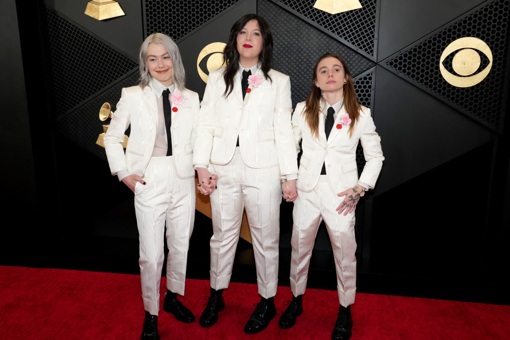 Phoebe Bridgers, Lucy Dacus, and Julien Baker on red carpet in matching white suits with pink corsages, holding hands at a music award event