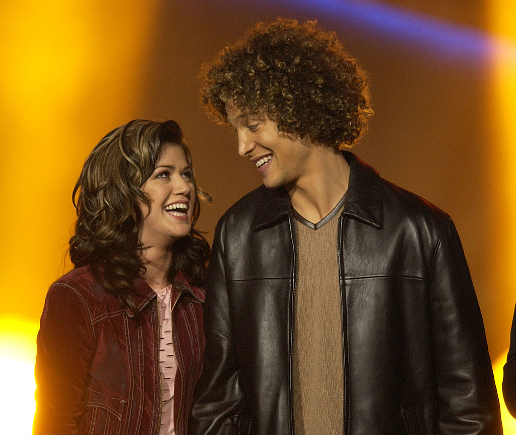 Kelly Clarkson and Justin Guarini smiling at each other on stage. Clarkson is wearing a casual jacket, and Guarini is dressed in a leather jacket