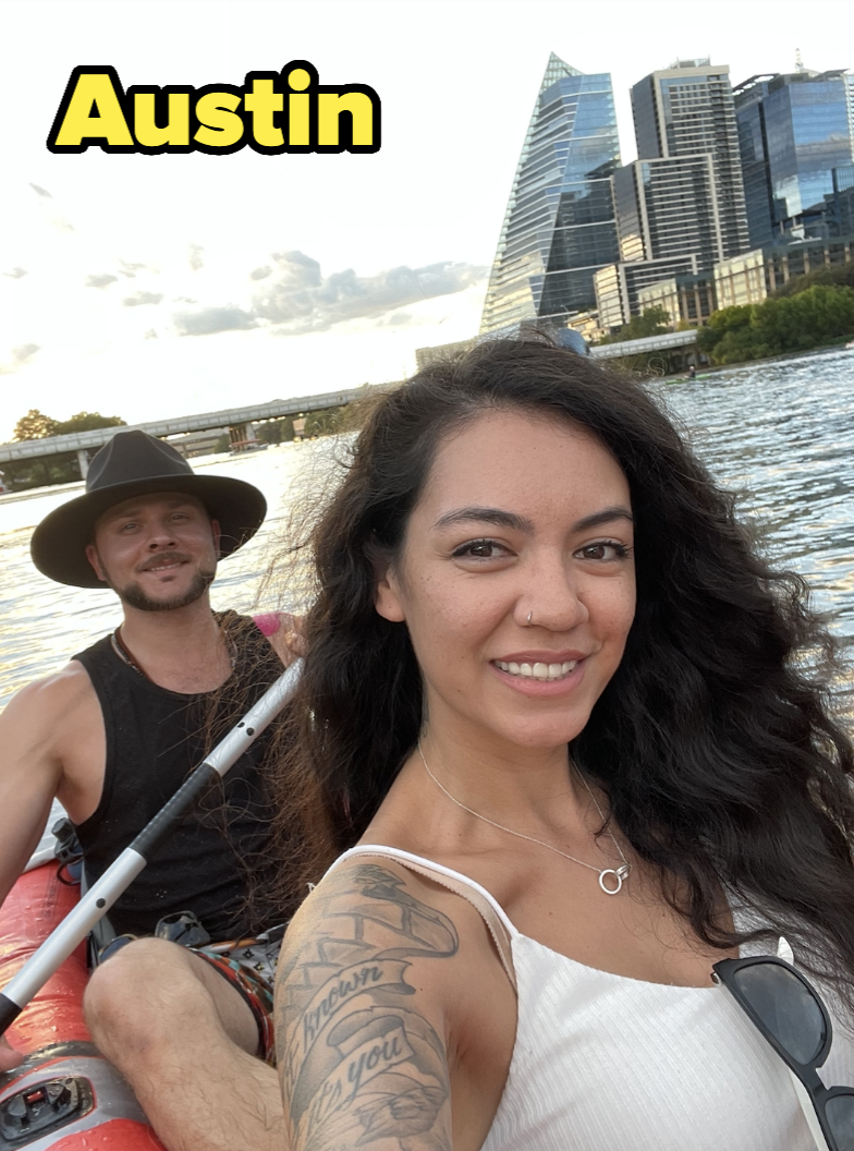 Man with a hat and woman with arm tattoos and a necklace kayaking in Austin with modern buildings in the background
