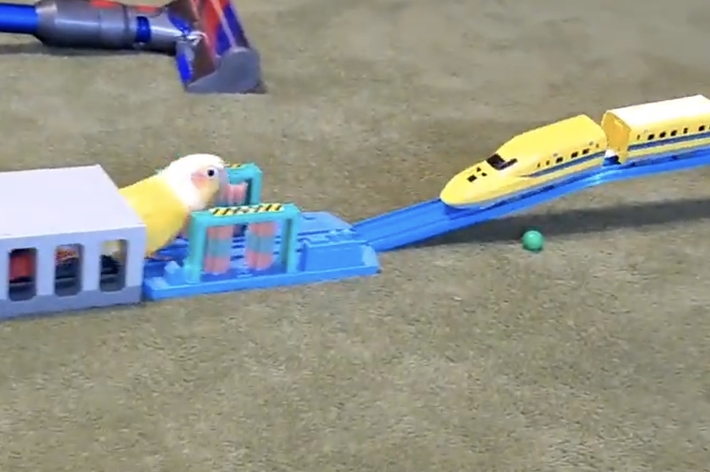 おもちゃの新幹線と黄色いパロット型のオブジェが、ミニチュアの鉄道セットで遊びます。広告記事用の画像です。