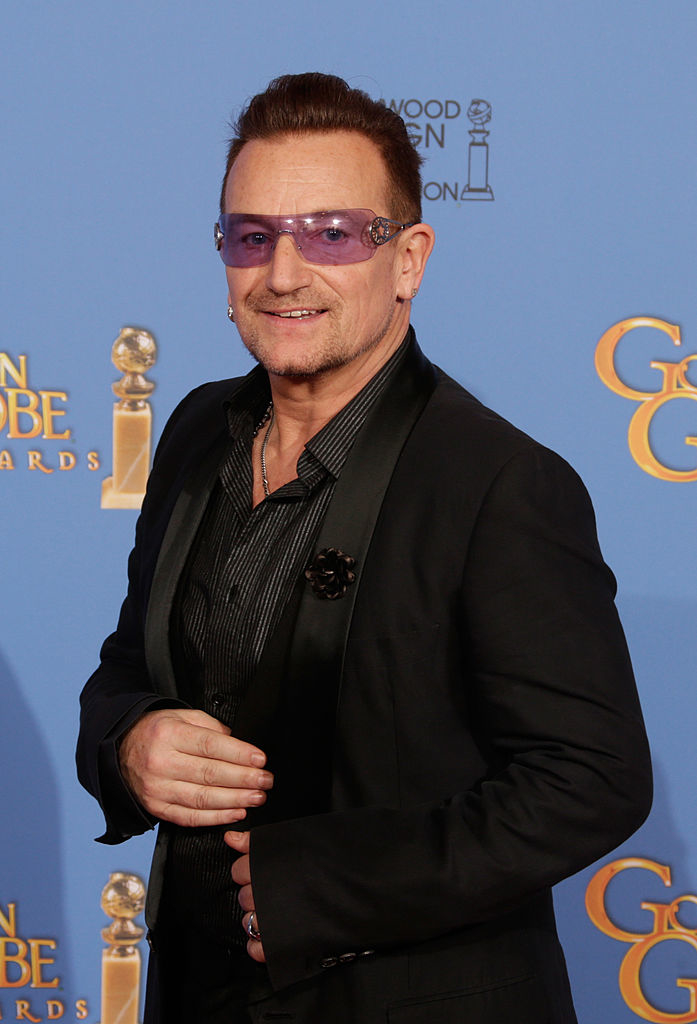 Bono wears a black suit with a striped shirt and purple sunglasses at an awards event