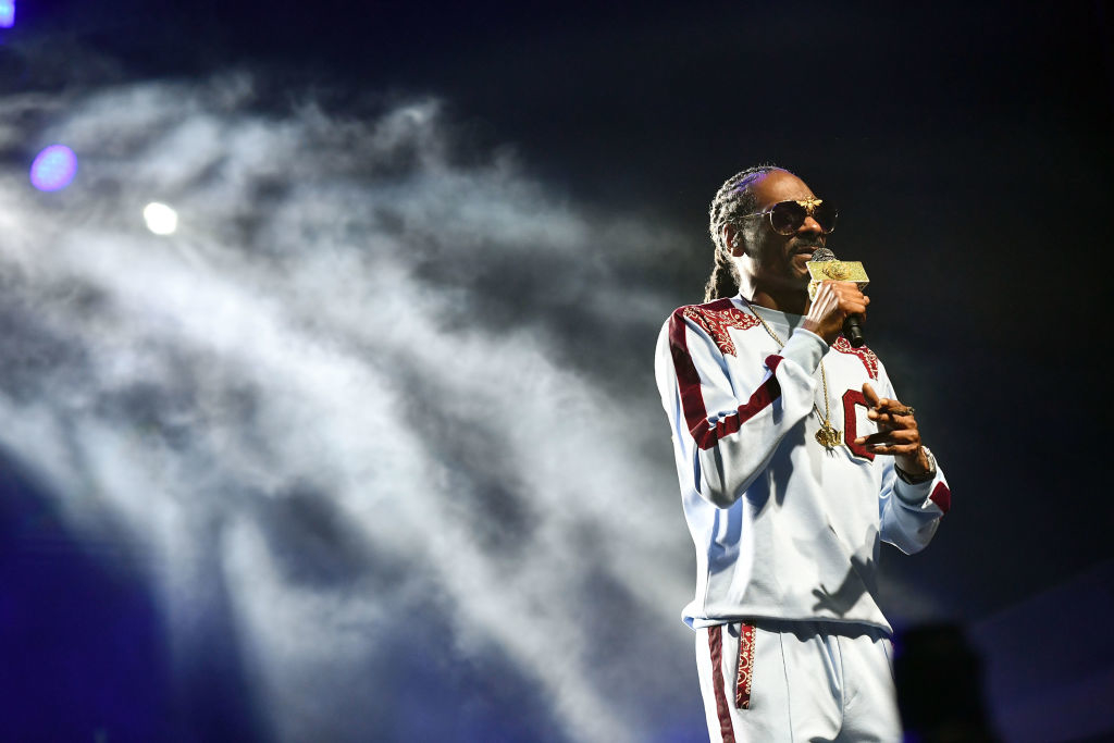 Snoop Dogg on stage, wearing a tracksuit, holding a microphone in one hand and a card in the other, with fog effects in the background