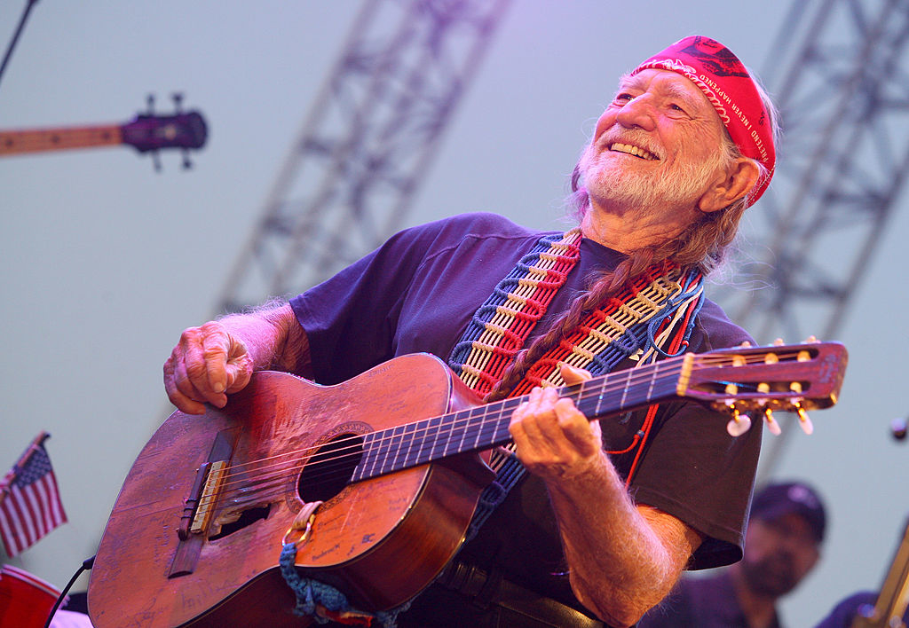 Willie Nelson performs on stage wearing a bandana and a beaded necklace, smiling while playing the guitar