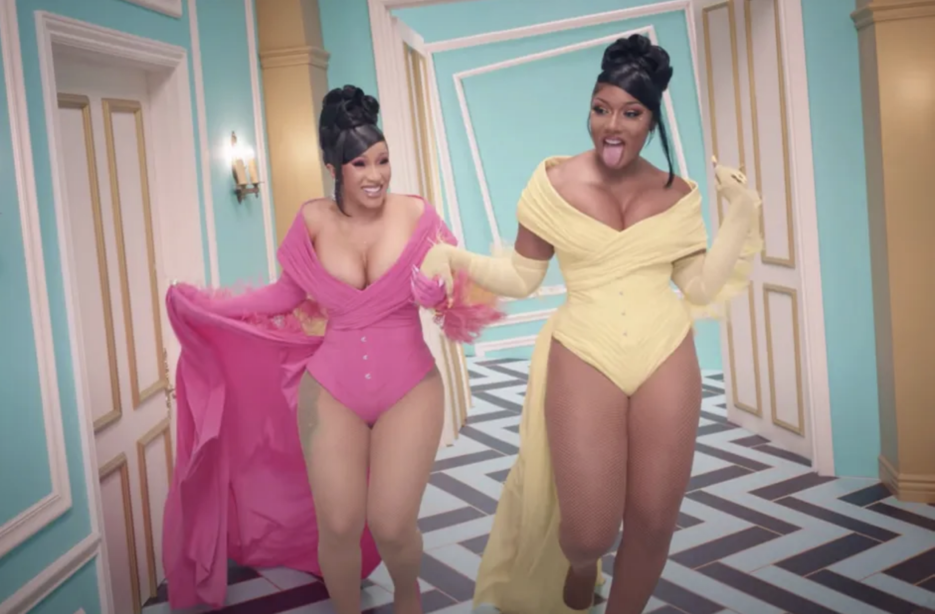Cardi B and Megan Thee Stallion in vibrant off-shoulder bodysuits, dancing in a dynamic hallway, from a music video scene
