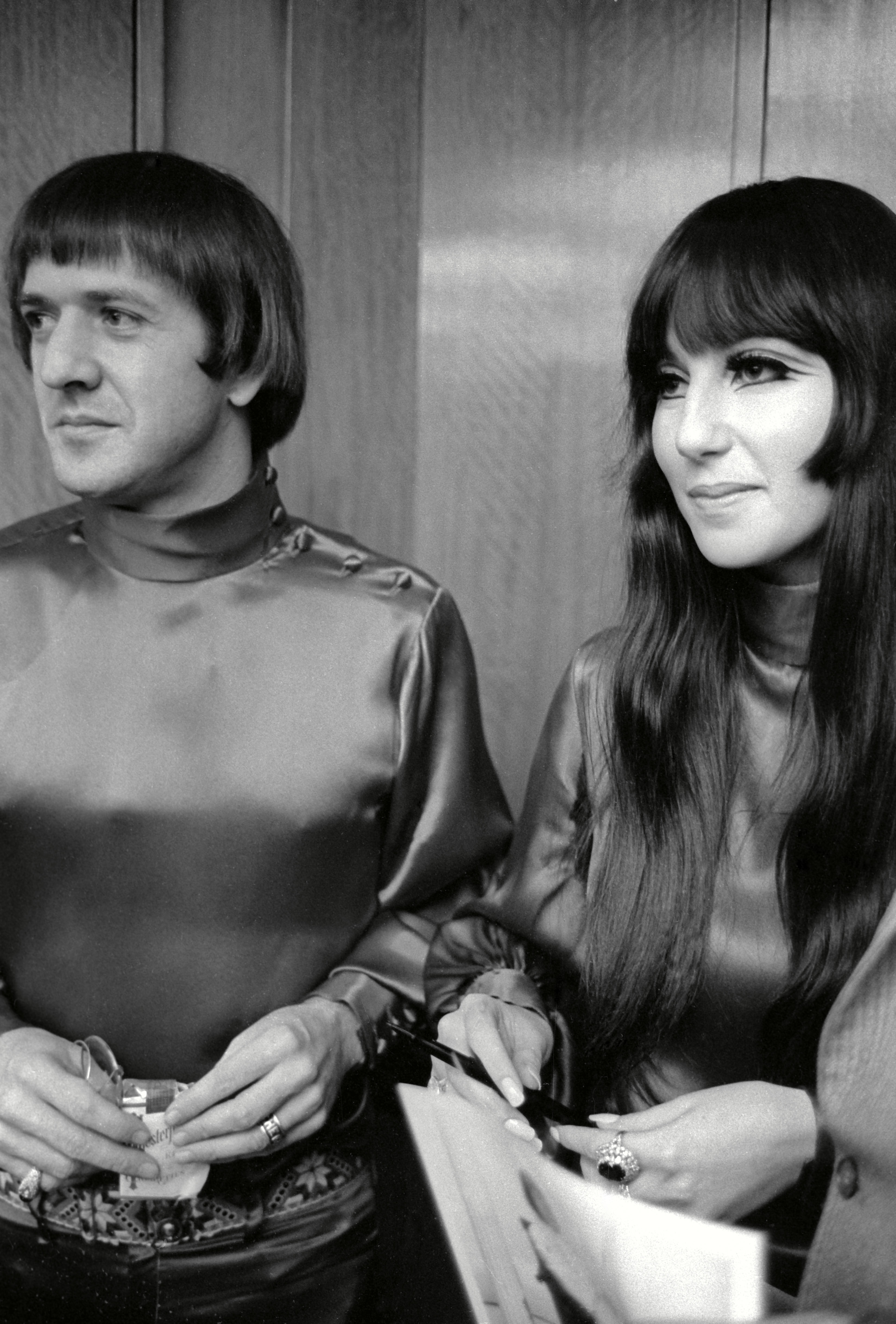 Sonny Bono and Cher in satin outfits, Sonny with a bowl cut hairstyle, and Cher with long, straight hair and bangs, both smiling while standing indoors