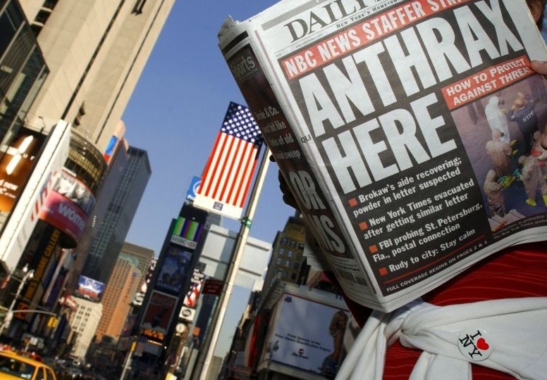 A person in Times Square reads a newspaper with the headline &quot;Anthrax Here.&quot; The article mentions NBC News and Tom Brokaw. Yellow taxis and tall buildings are visible